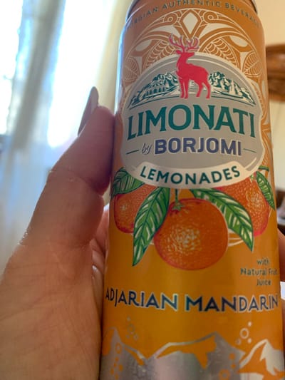 A close-up shot focuses on a hand holding a can of "Limonati by Borjomi Lemonades," specifically the "Adjarian Mandarin" flavor. The can has an orange background with intricate decorative patterns, a red deer silhouette against a mountain backdrop, and images of mandarins with leaves. The background of the image is blurred and appears to be indoors, with a window letting in bright, diffused light. The scene is not set in Mardakan, Azerbaijan, and there are no city elements visible.