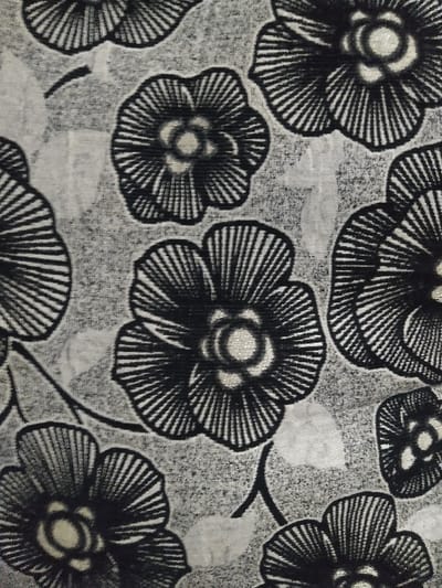 This image presents a detailed view of a textile or patterned surface, located in the city of Chimay, Belgium. The scene is dominated by a repeating pattern of stylized floral and botanical motifs rendered in a limited palette of black, white, and various grey tones. Multiple dark, five-petaled flowers are prominent, each distinguished by numerous thin, radiating lines forming the petals, emanating from a lighter, circular central core. These floral elements are interconnected and interspersed with less distinct, lighter grey, irregular leaf-like shapes and thin, curvilinear black lines suggestive of stems or vines. The background is a textured grey, providing a subtle visual depth to the pattern. No individuals, active subjects, or explicit interactions are present within the frame, which is entirely occupied by the patterned surface.