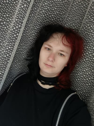 The image shows a young woman, likely in her late teens or early twenties, with black and red hair, wearing a black t-shirt and a black choker necklace. She appears to be lying down, possibly on a bed, as the background is a white lace fabric. The woman is looking directly at the camera with a neutral expression. The image appears to be a selfie taken indoors. The coordinates provided, 48.212, 22.64, correspond to a location in the city of Berehove, Ukraine, but this information is insufficient to determine the exact location of the image.