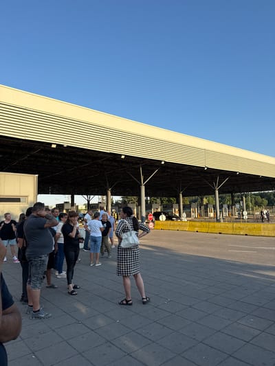 A group of people, a mix of men and women, are gathered on a paved area in front of a large, covered structure that appears to be a border crossing or customs checkpoint. The structure has a light-colored roof with horizontal slats and is supported by dark-colored pillars. Several customs booths are visible underneath the structure, with signs indicating "CUSTOMS". A yellow barrier runs along the road in front of the booths. In the background, there are trees and a clear blue sky. The people are casually dressed and some appear to be talking to each other, while others are looking forward or towards the customs booths. Some individuals are wearing t-shirts, shorts, and jeans, while others are in dresses or more formal attire. The scene appears to be outdoors during daylight hours. The image is taken from a slightly low angle, looking up towards the structure and the sky. The location is identified as Kapitan Andreevo, Bulgaria.