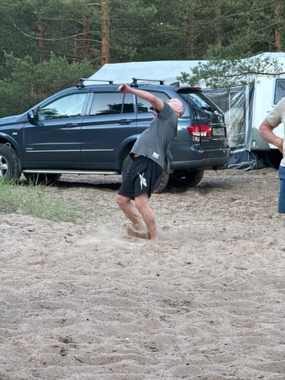 A man is mid-throw while playing a game in the sand at a campsite. He is wearing a grey t-shirt and black shorts. The man's body is arched backwards, and his right arm is extended upwards, having just released an object. Sand is kicked up around his feet, suggesting vigorous movement. Behind him, a dark grey SUV is parked on the sand, with a portion of a white caravan visible to the right. Trees form a backdrop to the scene. The image appears to be taken in Ozerki (Krasnodolinskaya), Russia, based on the provided context.