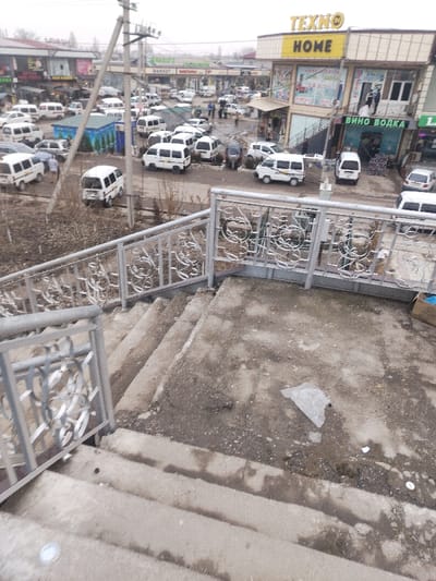 The image displays an elevated view of a commercial and transportation area in Navruz, Uzbekistan, under an overcast sky. In the foreground, concrete stairs descend, bordered by silver-colored metal railings featuring ornate, intertwined decorative patterns. The steps and an intermediate landing are visibly dirty, with scattered debris including a white plastic bag and small white fragments. A blue cardboard box is partially visible on the right side of the landing.
Below the stairs, a large, unpaved dirt and gravel expanse serves as a parking area, densely occupied by numerous white Chevrolet Damas mini-vans, with several individuals visible walking or standing among them. Sparse, bare vegetation protrudes from the ground. A utility pole with associated wiring stands prominently to the left of the center.
In the background, a row of commercial buildings lines the perimeter. A two-story building on the right features a prominent yellow and light brown facade with "TEXNO HOME" signage. Below it, a sign in Cyrillic reads "ВИНО ВОДКА" (Wine Vodka). To the left, another building displays a sign with "NAVRUZ MARKET" in both English and Cyrillillic. Additional indistinct figures are present near the buildings and vehicles.
