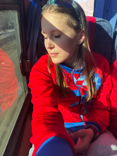 A young girl is seen resting, possibly sleeping, in what appears to be a vehicle. She is wearing a red and blue jacket and a blue headband. Her hand rests on a gray fabric. In the foreground, there is a glass surface, possibly a window or a screen, with a reflection of a red object. The location, Челва, Russia, is not evident in the visual details provided.