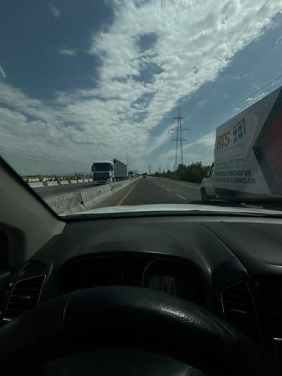 The image is taken from the interior of a vehicle looking out at a highway. The perspective is from the driver's seat, with the steering wheel and dashboard visible in the foreground. The highway is lined with concrete barriers and appears to have multiple lanes of traffic. Several trucks are visible on the road, including a white semi-trailer truck in the middle lane and another in the distance. A white van with "BTS" and text in a foreign language is parked on the right side of the road, partially obscuring the view of the highway ahead. Power lines and tall metal towers are present along the side of the road, indicating an electrical infrastructure. The sky is partly cloudy with a mix of blue sky and white clouds. The overall environment suggests a sunny day on a major road, possibly in an industrial or suburban area. The text on the van, "EXPRESS POCHTA VA LOGISTIKA XIZMATLARI," is in Uzbek and translates to "Express Post and Logistics Services." The location is identified as Istiqlol, Uzbekistan.