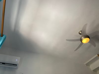 The image provides an upward-angled view of a room's interior ceiling and upper wall, located in Πλατύς Γιαλός, Greece. The ceiling is white. A brushed metal ceiling fan with three visible blades, illuminated by a warm yellow central light, is positioned in the upper right, its blades blurred due to rotation. In the lower left, a white, wall-mounted air conditioning unit is present, displaying the digital numeral "25" on its front panel. A light brown cylindrical wooden pole is partially visible extending vertically from the upper left, with a section of teal fabric draped below it. A white architectural molding or shelf is partially visible along the lower right edge of the frame.