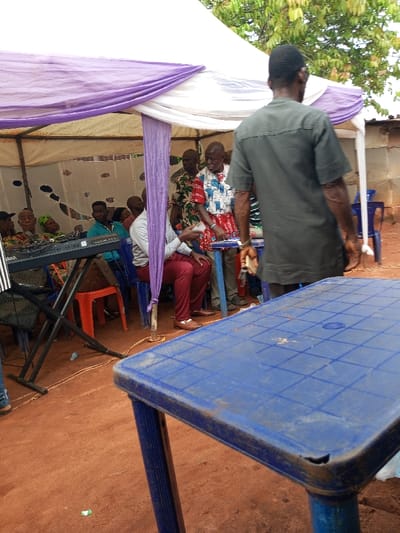 The image captures an outdoor scene in Oko Town, Nigeria, featuring a temporary gathering space under a white canopy tent with purple and white fabric drapes. The ground is composed of exposed red-brown earth, with minor debris including small plastic wrappers and orange cables visible.
In the immediate foreground, a weathered blue rectangular plastic table with a grid-patterned surface and a chipped corner is positioned. To its right, a male individual, observed from behind, stands wearing a dark green/grey short-sleeved tunic and a dark cap. He holds a white object in his right hand.
Within the tent, several individuals are present. On the left, a black Yamaha electronic keyboard rests on a black stand. Adjacent to it, a man in a light blue long-sleeved shirt is seated on a blue plastic chair. Towards the right side of the tent, an elderly man in a white and red patterned short-sleeved shirt stands near a small, light-colored table, holding a small item. Another man in a patterned shirt is visible standing behind him. To the left of the standing elderly man, a male in a light-colored shirt and red trousers is seated on a blue plastic chair, with his right arm resting on the small table. Additional individuals, including several elderly persons, are seated on blue plastic chairs and one red plastic stool further back within the tent. The tent structure is supported by metal poles. Beyond the tent's rear boundary, green foliage and a light-colored building are visible.