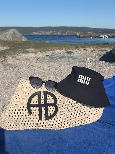 The image shows a collection of personal belongings on a beach in Nærsnes, Norway. A black bucket hat with a "Miu Miu" logo sits atop a woven beige bag adorned with a circular, abstract black logo. Sunglasses are positioned between the hat and bag. A blue towel is partly visible beside the bag. The beach surface is gravelly. In the background, the sea is visible under a clear blue sky. Along the coastline, there is a land formation with some vegetation.