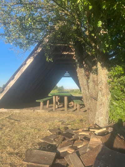 An outdoor scene in the village of Kozelshchyna, Ukraine, features a rustic wooden shelter with a triangular roof, open on one side to reveal a picnic table and benches painted green. The shelter is situated outdoors, with dry grass and a pile of chopped wood in the foreground. A large tree with a rough bark texture stands to the right of the shelter, its branches casting dappled shadows. The background shows a clear blue sky and distant greenery. The lighting suggests it is daytime.