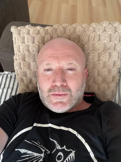 A middle-aged Caucasian man is lying on a sofa. The man has a shaved head, light stubble, and light-colored eyes. He wears a black t-shirt with a white graphic design. He is resting on a beige textured pillow. The sofa also has a striped pillow. The background includes a wooden floor. The image was likely taken indoors. There is no information in the image to indicate the location is Sint-Niklaas, Belgium.