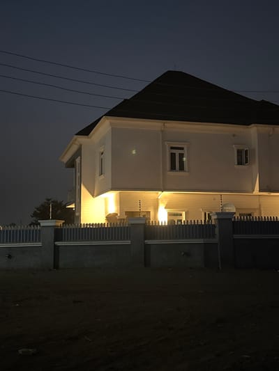 A low-angle, nocturnal view captures a two-story, light-colored building with a dark roof. Several windows are visible, some illuminated from within. The building's exterior is brightly lit, contrasting sharply with the dark surroundings. A fence borders the property. Power lines stretch across the upper left of the image. The night sky appears dark, and the ground is dimly lit, with minimal visible detail.