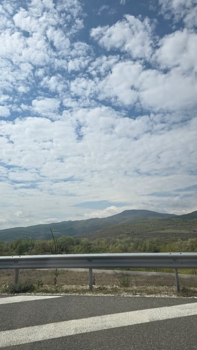 The image depicts a roadside scene with a view of mountains and a cloudy sky, likely taken on the outskirts of Valkovo, Bulgaria. The focus is on the edge of an asphalt road with a painted white lane marking and a metal guardrail. Beyond the guardrail, there is a grassy verge and a landscape of wooded hills, with mountains visible in the distance. The sky is predominantly blue, filled with numerous white clouds. There are no people visible.