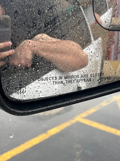 The image displays a close-up view of a wet passenger-side car mirror on a rainy day, located in the City of Middletown, United States of America. Numerous raindrops cover the mirror's surface, distorting the reflections. The mirror's black casing is visible along the bottom and left edge. Reflected within the mirror is a person's bare right arm, extending from the left, with the hand holding a black smartphone, suggesting the person is taking the photograph. A faint tattoo is visible on the upper arm. A white garment or object is partially reflected beneath the arm. The standard safety warning "OBJECTS IN MIRROR ARE CLOSER THAN THEY APPEAR" is printed on the lower part of the mirror's surface, partially obscured by raindrops and the reflection. The background reflection shows a dark, wet asphalt or concrete surface with distinct yellow painted lines, characteristic of a parking lot or street marking, indicating an outdoor environment during precipitation.