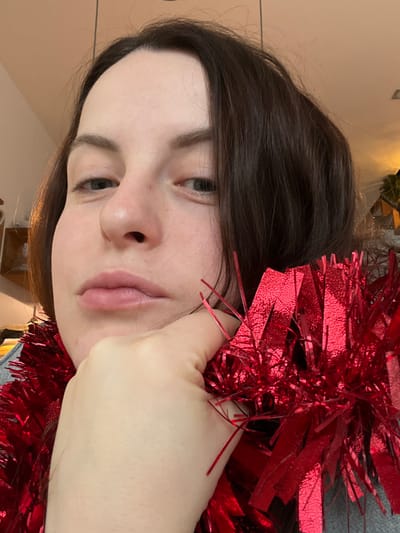 A female individual is depicted in a close-up, head-and-shoulders portrait, in an indoor setting within the city of Callbach, Germany. She has fair skin, dark brown hair, and light-colored eyes, and is looking directly forward with a neutral expression. Her left hand is raised, with her chin resting on the back of her fingers. A large quantity of bright red metallic tinsel or garland is draped around her neck and over her hand. The tinsel is composed of reflective, shimmering red strips. A portion of a grey garment is visible on her left shoulder. The background is blurred, showing indistinct indoor elements and diffuse lighting.