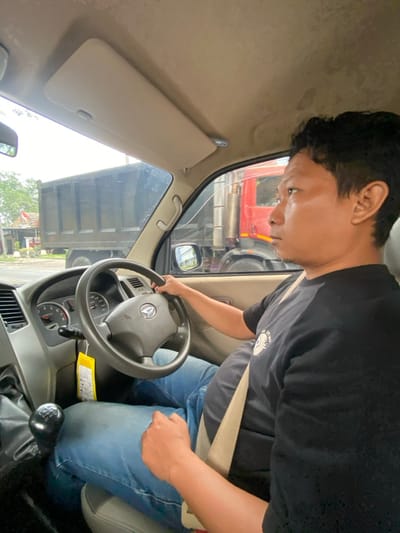 An Asian man wearing a black t-shirt and blue jeans is seated in the driver's seat of a truck. He has a beige seatbelt fastened across his chest. He is looking towards the windshield with his hands on the steering wheel. The interior of the truck is a light beige color. Through the windshield, a red truck is visible. The sun visor is down.