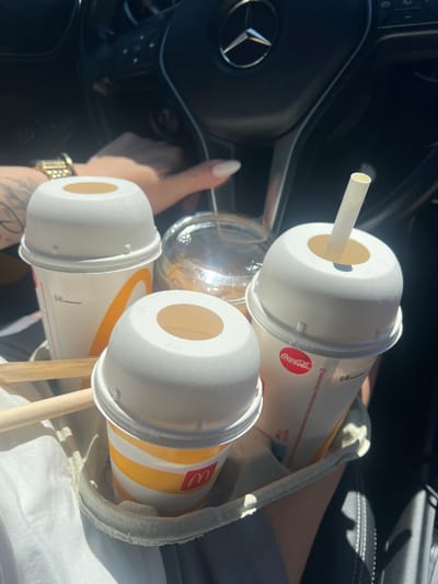 The image shows a person's hands holding multiple fast-food drinks inside a car. The scene is set in a car interior, with a steering wheel and part of the dashboard visible, suggesting the person is either driving or a passenger. The drinks appear to be from McDonald's and Coca-Cola, with their respective logos visible on the cups.

There are at least three large disposable cups with white lids and straws. Two of the cups are plain white with a Coca-Cola logo, and another cup has the distinctive yellow and red stripes of McDonald's packaging. One of the Coca-Cola cups has a straw inserted, and it's angled towards the viewer. A clear plastic cup, possibly with ice or a drink inside, is also visible.

The person's hands are prominent in the foreground. The right hand, with manicured nails, is resting on the steering wheel. The left hand appears to be holding or arranging the drinks. A watch with a gold band is visible on the left wrist. Tattoos are also visible on the left forearm.

The lighting suggests it is daytime, possibly sunny, as indicated by the bright highlights and shadows within the car. The overall impression is a casual snapshot of a person with drinks in a car, likely on the go.

Visible text includes "Coca-Cola" logo and "0.4L" on one of the cups. The McDonald's logo, the golden arches, is also visible on another cup.