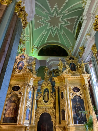 The image shows an ornate, gilded iconostasis within a church. The iconostasis is intricately carved and features several large paintings of religious figures, as well as smaller framed religious icons. Gold-plated statues of saints and angels are also present throughout the structure. The ceiling above the iconostasis is painted in a pale green color with a starburst design. The overall impression is one of grandeur and religious devotion. The scene is set inside a church in Ladozhskii Trudposelok, Russia.