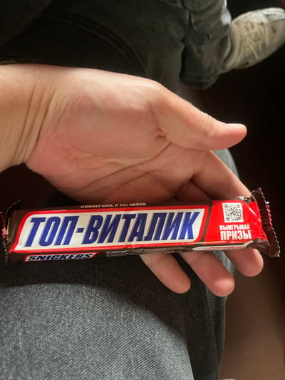 A hand is holding a Snickers chocolate bar. The wrapper is primarily red, white, and blue. The words "ТОП-ВИТАЛИК" are printed vertically in blue and white on the wrapper. The packaging also says "СНИКЕРСИИ, И ТЫ СПОВА", "На территории", and "ВЫИГРЫВАЙ ПРИЗЫ". A QR code is printed on the wrapper. The background shows a person wearing denim jeans and a shoe. The location appears to be indoors. There is no indication of location in the city of Красненскі сельскі Савет, Belarus.