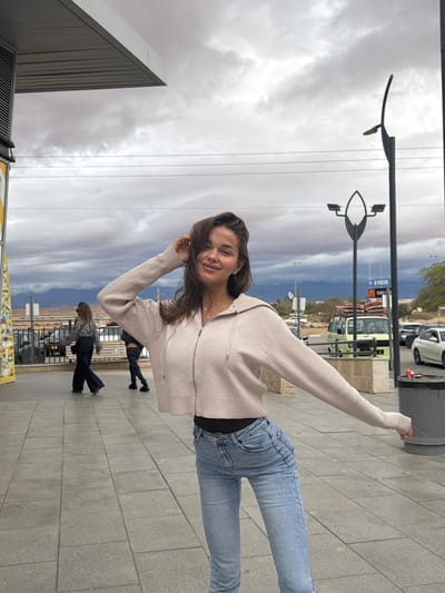 The image captures an outdoor street scene within the Arava Tichona Regional Council, Israel, under a heavily overcast sky filled with dark grey clouds. In the foreground, a young adult female stands facing the camera with a slight smile. She is wearing a light beige, ribbed, cropped zip-up hoodie over a black garment and light blue denim jeans. Her left arm is bent, with her hand positioned near her hair on the right side of her head, while her right arm is extended outwards. She wears a delicate silver necklace with a small clover-shaped pendant.
The ground consists of grey rectangular paving tiles. In the midground, two additional individuals are visible walking away from the camera. One person, with long hair, wears a patterned top and dark wide-leg pants, followed by another individual dressed in dark attire. Beyond the sidewalk, a low black fence delineates a parking area where several vehicles are present, including a light green Volkswagen Type 2 van with items on its roof rack, and various cars such as a white sedan.
Street furniture in the scene includes a tall black streetlamp with an elongated oval light fixture and another plain black pole. A blue sign featuring the McDonald's golden arches logo and Hebrew text is visible in the background. Distant reddish-brown mountains stretch across the horizon under the cloudy sky, indicating an arid landscape. A grey, perforated cylindrical trash can is located on the right side of the frame. An architectural overhang from a building is visible in the upper left corner.