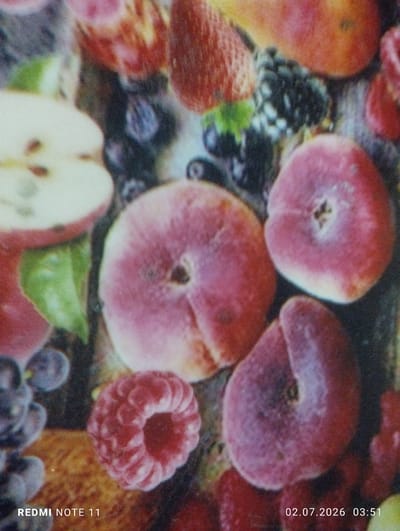 A close-up, overhead shot shows a collection of ripe fruits, possibly a still life or part of a pattern on a surface. The fruits are arranged closely together and appear to be a mix of berries, peaches or nectarines, grapes, and what looks like a sliced apple. A raspberry is prominent in the lower-left quadrant, and a flat, circular peach or nectarine is centered in the frame, with a darker, oval-shaped fruit to its right. Grapes, some appearing as dark purple berries, are visible to the left and in the upper portion of the image. Other red fruits, potentially strawberries or cherries, are also present, creating a vibrant, colorful composition. The lighting is diffused, highlighting the texture and gloss of the fruits. Text overlays at the bottom of the image read "REDMI NOTE 11" on the left and "02.07.2026 03:51" on the right. There are no people, animals, or discernible environmental features that would indicate a specific location within the City of Türkmenabat, Turkmenistan.