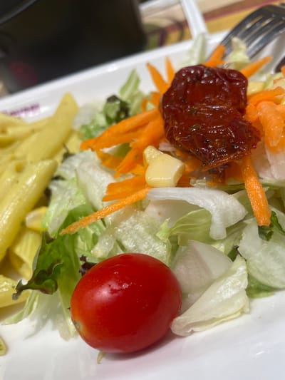 This is a close-up, top-down shot of a meal served in a white bowl. The meal appears to be a combination of pasta and salad. The pasta, predominantly in the foreground, consists of penne tubes coated in a creamy yellow sauce. The salad, taking up the upper portion of the frame, is a mix of shredded lettuce, shredded carrots, a single cherry tomato, and what looks like a sun-dried tomato. There are also a few small pieces of what might be corn or cheese scattered throughout the salad. A fork, with its tines visible, is positioned on the left side of the frame, suggesting the meal is ready to be eaten. The lighting is bright, hinting at daytime. The overall impression is a fresh and appealing dish. No text is visible in the image. The location context of Bağcılar, Turkey is provided but not directly evident from the image itself.