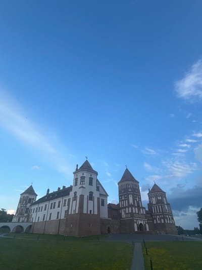 The image displays the Mir Castle Complex, a UNESCO World Heritage site located in the village of Mir, Korelichsky District, Grodno Region, Belarus. The castle, a striking example of Gothic, Renaissance, and Baroque architecture, is viewed from a low angle, emphasizing its imposing structure against a clear blue sky with wispy clouds. The foreground features a well-maintained green lawn, with a paved pathway leading towards the castle. The castle itself is characterized by its distinct white and red brick walls and multiple towers topped with conical roofs. A bridge with arched supports is visible on the left side, connecting to the castle complex. The scene depicts the castle during daytime, under a bright sky, suggesting a pleasant weather day. No people or animals are discernible in the image.