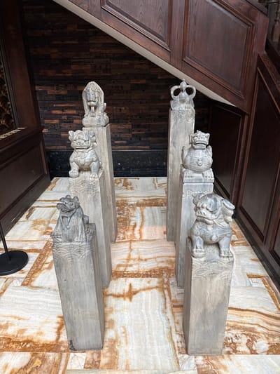 The image displays an indoor scene in Tongxiang, China, featuring six grey stone sculptures positioned on six rectangular stone pillars. The sculptures, arranged in two staggered rows of three, depict stylized mythological creatures. From left to right in the foreground, there is a seated frog-like creature and a seated foo dog/lion. In the middle ground, there is a standing foo dog/lion with an open mouth and a crowned, bird-like mythological figure. In the background, a seated foo dog/lion and a turtle-like creature with a patterned shell are visible. Each pillar has a decorative wave-patterned carving below its respective sculpture.

The floor is composed of highly polished, cream and light brown marble tiles with darker brown veining, arranged in a grid pattern. The background features dark brown wooden wall paneling with horizontal plank details. On the right, a dark brown wooden staircase with carved railings and raised panels ascends out of frame. A black, circular base on a stand is partially visible on the far left. No individuals are present in the scene.