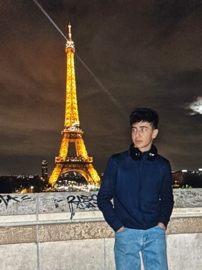 A young male stands in the foreground, positioned slightly right of center, against a nighttime cityscape. He has dark, curly hair and wears a dark blue long-sleeved zipped jacket, light blue denim jeans, and black over-ear headphones resting around his neck. His hands are in his pockets, and he gazes towards the right side of the frame. Behind him, the iconic Eiffel Tower is prominently illuminated with golden lights, featuring a visible light beam extending diagonally upwards from its spire into the dark, cloudy sky. A distant urban skyline with various scattered lights is visible beneath the tower, along with another tall building to the left of the tower's base. In the immediate foreground, a weathered stone balustrade or wall, covered with numerous scribbled markings and graffiti, acts as a barrier. This scene is located in the city of Dehqonbozor, Uzbekistan.
