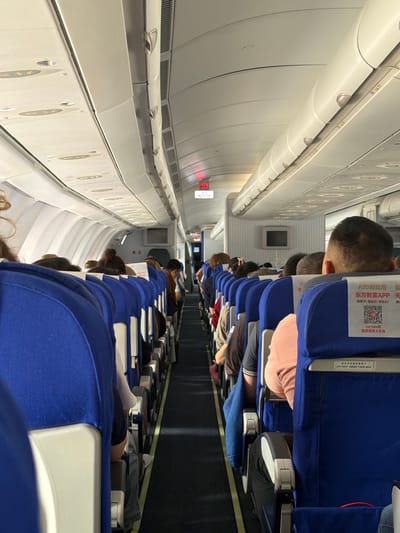 The image shows the interior of a commercial airplane cabin, with rows of blue seats occupied by passengers. The perspective is from the aisle, looking forward. The overhead compartments and lighting fixtures are visible along the top of the cabin. An illuminated exit sign is visible in the distance. On the back of the seat in the foreground, there is a safety card with Chinese text and a QR code. The scene is indoors and appears to be in flight. The location in Guangyang District, China is not discernible from the image.