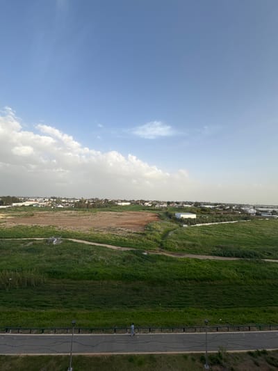 An elevated long shot depicts a semi-urban landscape in Yavne, Israel, under a partly cloudy blue sky. The upper half of the image features a clear blue sky transitioning to scattered white cumulus clouds along the horizon line, particularly prominent on the left. The midground consists of an expanse of mixed terrain: a large, irregular brown field occupies the central-left, bordered by vibrant green fields and vegetation to its right and foreground. Dirt paths traverse these green areas, and a small, white, possibly utilitarian structure is visible within the green field in the center-left. Further in the background, a density of residential and industrial buildings, interspersed with trees, defines a cityscape along the horizon. A white-roofed building is present in the mid-right within a green section. In the foreground, a dark asphalt road or pathway stretches horizontally across the bottom of the frame, separated from the green fields by a dark, low-height guardrail. Cracks are visible on the road surface. A single individual, dressed in light-colored clothing, walks from right to left on this paved path. A dark grey streetlamp pole stands upright on the right side of the path, extending upwards from the lower frame edge.