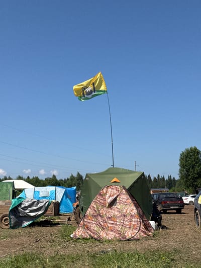 A campsite is set up on a grassy field under a clear blue sky. A yellow and green flag with a symbol on it waves from a pole in the center of the frame. Several tents are visible, including a large green tent, a camouflage-patterned tent, and other tents in the background. A trailer covered with a tarp is positioned to the left of the tents. Cars are parked in the background. The ground is uneven and appears to be dirt and grass. The overall impression is of an outdoor gathering or festival. The scene appears to be in the city of Chyormoz, Russia, judging by the context provided.