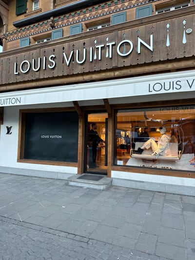 The image depicts the exterior of a Louis Vuitton store in Gstaad, Switzerland. The building features traditional Swiss chalet architecture with dark wooden paneling, white 'LOUIS VUITTON' lettering prominently displayed above a white awning, and upper-story windows fitted with dark green wooden shutters. Stylized tie-shaped wooden carvings adorn the facade above the main sign. The storefront comprises two large display windows flanking a recessed central entrance with a dark mat and stone step. The left display window is largely covered by a black panel featuring the 'LOUIS VUITTON' logo in white, with a smaller black 'LV' logo visible on the white wall segment to its left. The right display window is transparent, showcasing an interior scene where a mannequin, dressed in a light-colored patterned ski suit, dark boots, and goggles, is seated within a white ski lift chair. Other store interior elements and additional mannequins are visible behind the primary display. A male individual, dressed in dark professional attire, is visible inside the store near the entrance, facing towards the right interior. The sidewalk in the foreground is paved with grey stone or concrete tiles, exhibiting minor surface variations.