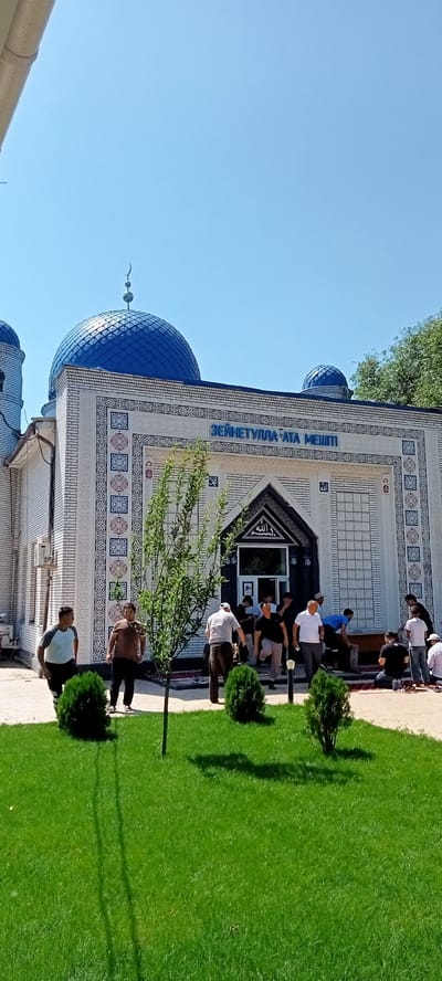 This is a full-shot, eye-level, outdoor photograph of a mosque in Besagash, Kazakhstan. The mosque features white brickwork with decorative blue and red tile patterns. It has prominent blue domes and is topped with a crescent moon finial. The text above the entrance, "ЗЕЙНЕТУЛЛА АТА МЕШІТ," is visible.
In the foreground, a well-maintained green lawn with manicured bushes surrounds a young tree. Several men are gathered in front of the mosque entrance, some standing and some kneeling on prayer rugs. The men are dressed in casual attire, and the overall scene suggests a daytime gathering, possibly for prayer. The sky is clear and blue.