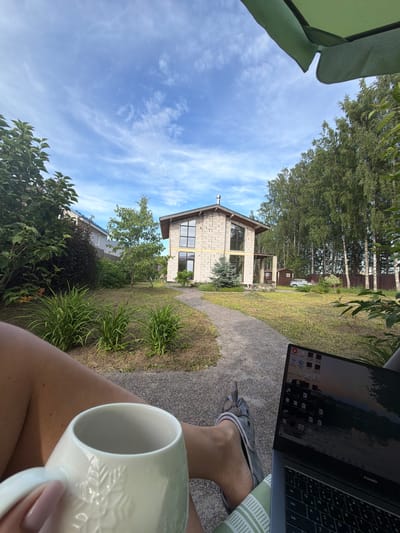 The image shows a first-person perspective of someone sitting outdoors in a garden with a laptop and a mug. In the background, there is a modern, multi-story house constructed with light-colored bricks, featuring large windows. The garden area is characterized by green grass, some ornamental plants, and a pathway leading towards the house. Tall trees, primarily birch trees, form a dense line along the right side of the property. A fence is visible behind the trees. The sky is blue with scattered white clouds. A silver car is parked in the driveway near a small shed. The person is wearing casual clothing, including a sandal, and is holding a white mug. The scene appears to be a residential area, possibly a dacha or a suburban home. The location is identified as Kerro, Russia.