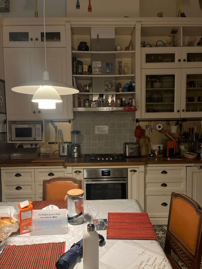 This image displays a well-lit kitchen and dining area in Saint-Gilles - Sint-Gillis, Belgium.
In the foreground, a dining table is covered with a white tablecloth. Upon it are two rectangular placemats with a textured red pattern. Items arranged on the table include a clear glass pitcher with a cork lid holding water, a white cylindrical water bottle, a dark blue folded umbrella, and a white upright sign. This sign features black text that reads, "The Kitchen space is OPEN from 8AM till 10PM," followed by additional informational text. To the left, an orange carton box and a plastic bag containing baked goods are partially visible. A brown-orange upholstered chair with a carved wooden frame is positioned at the table, partially obscured.
The background reveals a kitchen equipped with cream-colored cabinetry, a dark wooden countertop, and a grey-and-white patterned tiled backsplash. A large, multi-tiered white pendant light fixture hangs from the ceiling, centrally illuminating the space. A white sign affixed to the backsplash states "Only Veg Food." Kitchen appliances visible on the countertop include a silver microwave oven on a shelf to the far left, a silver toaster, and a silver blender. A four-burner gas hob is integrated into the countertop, directly above a stainless steel built-in oven whose digital display reads "12:00." To the right, various kitchen utensils, cutting boards, and a red espresso machine are arranged on the countertop. Above the countertop, upper cabinetry consists of both closed white doors, open shelving units displaying decorative items and spices, and glass-fronted cabinets revealing stacked plates and bowls. No individuals are present in the scene.