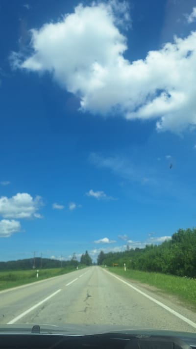 The image captures a scenic view of a highway in Glāžšķūnis, Latvia, on a bright, sunny day. The sky is a vibrant blue, adorned with fluffy white clouds. The road stretches out into the distance, with white lane markings guiding the way. Lush green fields and dense forests flank both sides of the highway, creating a picturesque landscape. The perspective suggests the photo was taken from a moving vehicle, likely a car, looking forward. There are no people or animals visible in the frame. The overall atmosphere is one of a peaceful and scenic drive through the Latvian countryside.