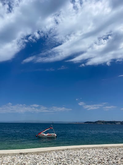 This is a landscape-oriented photograph taken at Crveni Vrh, Croatia. The image captures a vibrant blue sky filled with wispy white clouds, with the horizon meeting a deep blue sea. Along the right edge of the frame, a shore composed of small, light-colored pebbles is visible, separated from the water by a concrete border.

In the water, a red and white pedal boat is present, its form partially submerged. It appears to be a recreational device, possibly for rent or personal use. The water is calm, with a few small buoys marking a perimeter in the distance. Beyond the sea, a coastline dotted with buildings and greenery stretches across the horizon, suggesting a populated area. The overall scene evokes a sense of a clear, sunny day, ideal for outdoor activities by the water. The bright lighting and the clarity of the sky suggest it is likely daytime, perhaps late morning or afternoon. There is no visible text in the image.