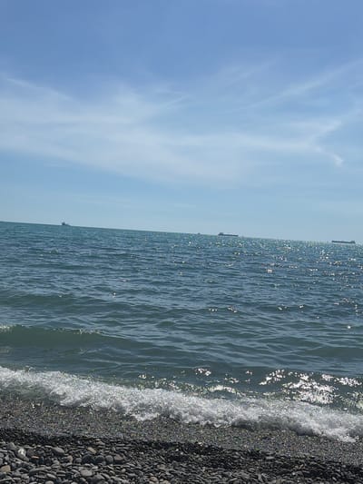 A wide, sunny shot shows a pebble beach leading to a calm, blue sea. The water is sparkling with sunlight, and gentle waves lap at the shore. In the distance, several large ships are visible on the horizon. The sky is a pale blue with wispy white clouds. The scene evokes a peaceful and serene atmosphere, characteristic of a seaside location in Shirokaya Balka, Russia.