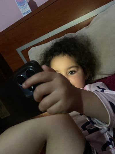 A child is lying down, possibly on a bed or a soft surface. The child has dark curly hair and is holding a smartphone in front of their face. The phone is positioned as if taking a selfie or looking at the screen. The background shows parts of a room, including a wooden surface and possibly medication packaging. The image location is specified as Sidi Slimane Moul Al Kifane, Morocco.