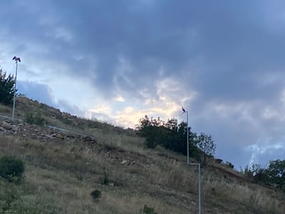 The image depicts a hillside landscape in Balgarchevo, Bulgaria, under an overcast sky with patches of brighter light visible through the clouds, suggesting a potential time around sunrise or sunset. The hillside is covered with dry, sparse grass and scattered rocks, indicating arid conditions. Several small bushes and trees are visible across the slope. Two tall, slender poles rise from the hillside, each topped with a distinct, reddish-purple object. The object on the left pole appears somewhat winged or folded, while the one on the right pole is more singular and elongated. No people or animals are visible, and no specific actions are occurring within the frame. The overall scene is static, focusing on the natural terrain and the two artificial structures against the cloudy sky.