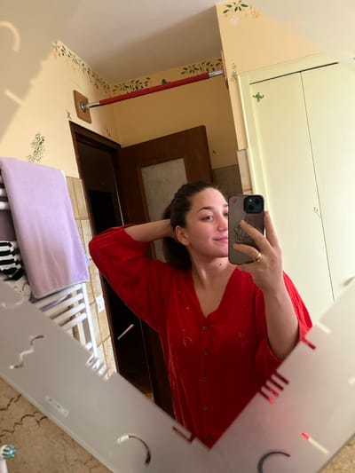 The scene depicts a mirror selfie taken in a domestic bathroom located in Ancenis, France. A female with medium-length dark hair, pulled back, is visible in the reflection. She is wearing a red, collared, long-sleeved blouse featuring a button-front closure and eyelet embroidery. Her left hand is positioned near the back of her head, while her right hand holds a dark-cased smartphone, used to capture the image. A gold-colored ring is present on her left ring finger. The mirror has an irregular, decorative shape with frosted or etched cut-outs along its lower and left edges. Reflected in the mirror, the bathroom walls are painted pale yellow with green and beige floral stencil patterns. A white ladder-style towel rack, holding a light purple towel and a black-and-white striped towel, is visible on the left. Above it, a red-covered metal bar, secured by a wooden mount, extends into the corner. A dark wooden door frame is partially visible in the background. To the right, a light-colored two-door cabinet, also adorned with small green stencil designs, is reflected.