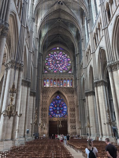This is an interior shot of a large Gothic cathedral, specifically the Reims Cathedral, located in the city of Reims, France, not Urvillers. The perspective is from the nave, looking towards the altar and the stained-glass rose windows. Massive stone columns with intricate carvings line both sides of the nave, supporting ribbed vaulted ceilings. Two large, ornate stained-glass rose windows, one above the other, dominate the far wall, displaying a vibrant array of colors. Below the upper rose window are rectangular stained-glass panels. Rows of empty wooden chairs are set up in the nave, with a few people walking in the background near the altar area. The overall impression is one of grandeur and historical significance.