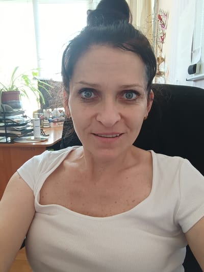 A woman with dark hair pulled up in a bun and blue eyes is looking at the camera. She is wearing a white ribbed shirt with a cutout neckline. She has a light complexion and freckles. The background appears to be an office or home setting, with a desk to her left holding office supplies, plants, and a bottle of lotion. A window with blinds is visible behind the plants, and a black office chair is behind her to the right. The image is a close-up selfie. The location of Dnestrovsk, Moldova is not visually discernible from the image.