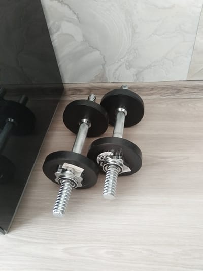 Two dumbbells are placed on a wooden floor. The dumbbells have black plates and chrome handles with a ribbed grip. One dumbbell is in the foreground on the right, and the other is slightly behind and to the left. A black, reflective surface, possibly a piece of furniture or appliance, is visible on the left side of the image, reflecting the dumbbells. The background features a tiled wall with a marbled pattern in light gray and white. The overall setting appears to be an indoor space, possibly a home gym or a room with exercise equipment. There are no people present in the image.