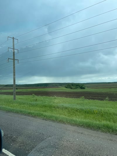 The image depicts a rural scene, likely in the vicinity of Кокино, Russia, based on the prompt. A utility pole and associated power lines dominate the left side of the frame against a cloudy sky. A field of cultivated land is visible on the right, interspersed with grassy areas and possibly some vegetation. A road runs alongside the field. The weather appears overcast, suggesting possible precipitation. No people are visible in the image.