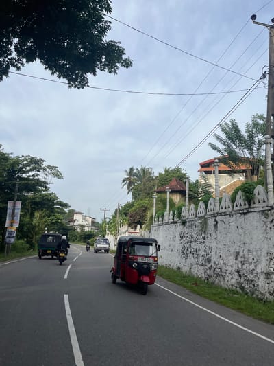 In Walahanduwa, Sri Lanka, a roadside scene unfolds under an overcast sky. A paved road, marked with a white dashed line, curves through a lush green environment. On the right side of the road, a textured white wall with decorative triangular cutouts runs alongside, topped with a railing. Behind the wall, a multi-story building with a red-tiled roof is visible amidst dense foliage.
Several vehicles navigate the road. A red three-wheeled auto-rickshaw, displaying the license plate "SB ABJ 5891," occupies the foreground on the right. Further down the road, a white van is in motion. A person on a motorcycle, wearing a white helmet, travels ahead of the van. On the left side of the road, a dark green three-wheeled auto-rickshaw is also moving. Alongside the road on the left, a tall pole supports multiple power lines that crisscross the sky. A billboard with text in a local script and English is affixed to a pole on the far left. Trees and vegetation line both sides of the road, with palm trees adding to the tropical atmosphere. The overall scene suggests a typical day on a road in this Sri Lankan locality.