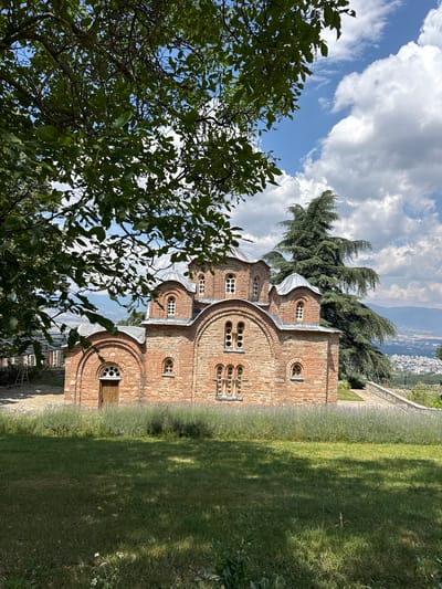 This media file captures an outdoor scene featuring a historic brick church. The church has a distinctive architectural style with multiple domed roofs and arched windows, some of which contain multiple small circular openings. The brickwork is weathered and shows a variety of tones, suggesting age.

In the foreground, lush green foliage from a large tree partially obscures the view of the church. To the right of the church, a well-maintained lawn slopes gently downwards, with dappled sunlight creating patterns of light and shadow. Beyond the church and lawn, a distant cityscape and rolling hills are visible under a partly cloudy sky. The clouds are dramatic and white, contrasting with the bright blue sky. The overall impression is one of historical significance and natural beauty. The image is taken from a slightly elevated viewpoint, looking down towards the cityscape. There is no visible text in the image. The lighting suggests it is daytime, likely mid-morning or afternoon, given the shadows. The weather appears pleasant, with a mix of sun and clouds.