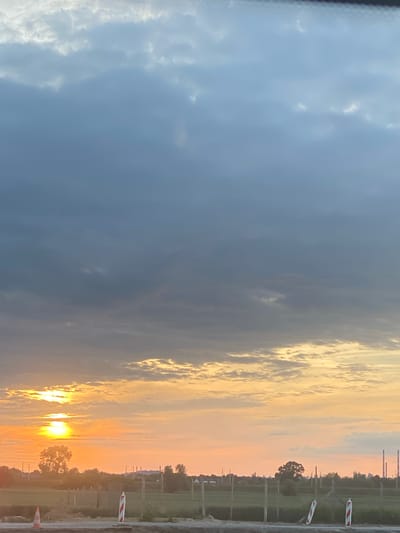 The image depicts a landscape at dusk, likely in Podłęże, Poland. A vibrant sunset casts an orange and yellow glow across the sky, illuminating scattered clouds. In the foreground, a construction zone is visible, with a safety cone and striped bollards. Beyond the construction site, a field stretches towards a treeline, with several industrial smokestacks in the distance. The scene is devoid of people.