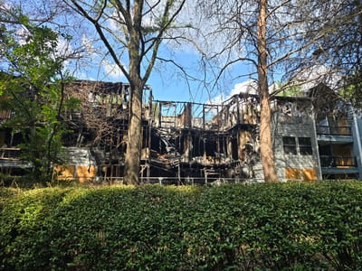 In Roswell, United States of America, a heavily fire-damaged apartment building stands partially obscured by trees and a thick green hedge. The central section of the building is completely gutted, with charred wooden beams and debris exposed to the sky. The exterior walls of the remaining sections are clad in light-colored siding, with some windows intact and others boarded up with plywood. A white metal fence runs along the front of the property, separating the hedge from the damaged structure. The sky is blue with scattered white clouds, and the sun is casting shadows.