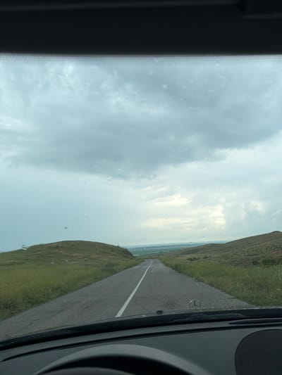 The image, which appears to be rotated 90 degrees clockwise from its natural orientation, depicts a view from inside a vehicle traveling on a road. When correctly oriented, the asphalt road stretches ahead, disappearing into the distant horizon, marked by a single white line on its right edge.

On either side of the road, gently sloping hillsides are covered in lush green grass, suggesting a rural or open landscape. The sky above is largely overcast with thick, grey clouds, indicating a cloudy or partly cloudy day. Small specks, possibly water droplets or dust, are visible on the vehicle's windshield.

Parts of the car's interior frame the scene on the left and right edges. On the right side of the frame (which would be the top right if rotated), a dark dashboard or pillar is visible. A prominent Mercedes-Benz logo, a three-pointed star within a circle, is seen on the car's interior near the windshield, indicating the brand of the vehicle. The scene captures a moment of travel or driving through this open, natural environment. Based on the provided context, this location is Kovachite, Bulgaria.