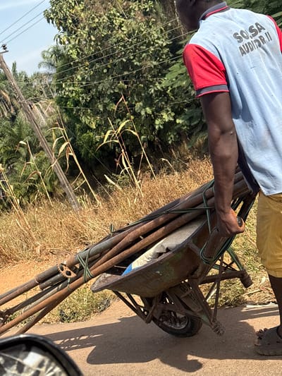 The scene, located in Tataura, Nigeria, depicts a male individual pushing a heavily laden wheelbarrow on a dark gray asphalt road under bright daylight. The man, observed from the rear-right, wears a light blue short-sleeved shirt with red shoulder panels and collar trim, displaying the text "SO, SA AUDITOR II" on the back. He also wears yellow shorts and dark open-toed sandals. His right arm is visible, actively gripping the right handle of the wheelbarrow.
The wheelbarrow is a single-wheel model, green in color with visible rust. It contains multiple long, cylindrical, rusty metal pipes or poles, secured together by green strapping. Beneath these poles, a light-colored bag, possibly containing cement or a similar material, rests within the wheelbarrow's basin.
The immediate roadside consists of dry, tall, light-brown grass and sparse green vegetation. Further into the background, dense green foliage, including palm trees, and utility poles with multiple overhead wires are visible against a clear, light blue sky. A partial view of a reflective surface, consistent with a vehicle mirror, is present in the lower-left foreground. Strong shadows are cast on the road, indicating direct sunlight from above and to the left.